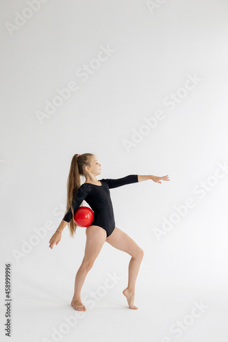 slim artistic teenager girl in black leotard trains on white background with red ball in her hands in rhythmic gymnastic exercise, children's professional sports