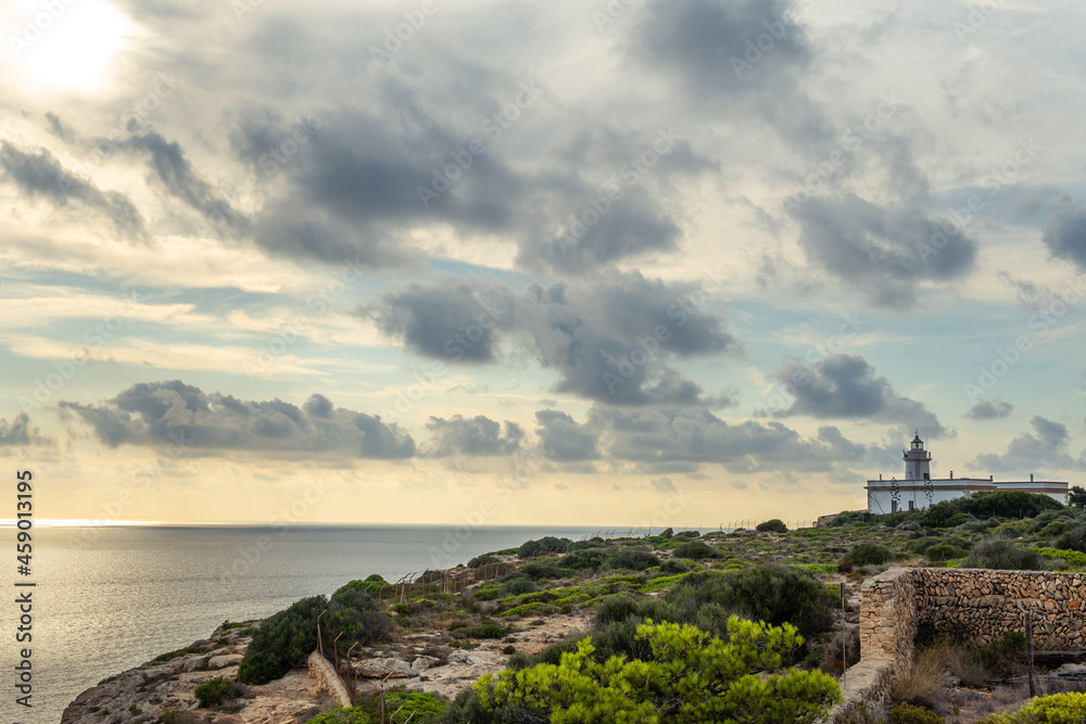 Cap Blanc lighthouse on the rocky coast of the island of Mallorca. In the background s seascape of the Mediterranean Sea at sunset with clouds. Sublime landscape