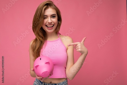 Portrait photo of sincere happy positive smiling sexy young charming woman with light hair wearing stylish pink top isolated over pink background with empty space and holding pink pig penny bank