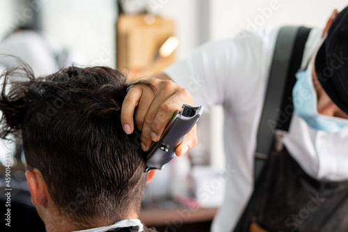 Barber with face mask using hair clipper on client. Stylist cutting hair with machine.