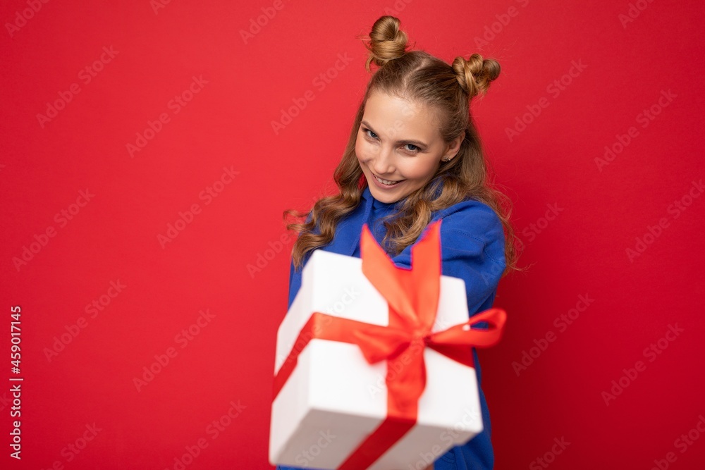 Charming happy funny joyful young blonde woman isolated over red background wall wearing blue trendy hoodie holding gift box and looking at camera Charming happy funny joyful young blonde woman isolated over red background wall wearing blue trendy hoodie holding gift box and looking at camera