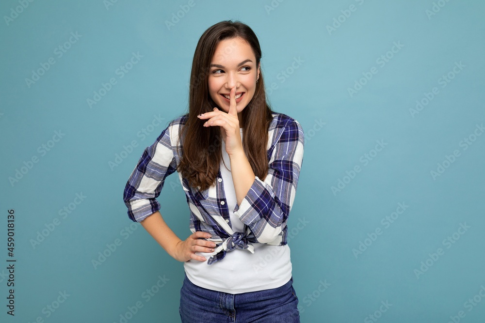 Young positive smiling beautiful brunet woman with sincere emotions wearing trendy check shirt standing isolated on blue background with empty space and showing shh gesture Young positive smiling beautiful brunet woman with sincere emotions wearing trendy check shirt standing isolated on blue background with empty space and showing shh gesture