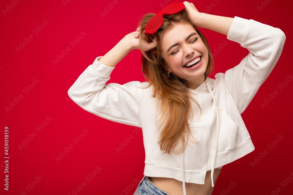 Shot of beautiful happy smiling young blonde woman isolated over red background wearing casual white hoodie and stylish sunglasses enjoying and having fun Shot of beautiful happy smiling young blonde woman isolated over red background wearing casual white hoodie and stylish sunglasses enjoying and having fun