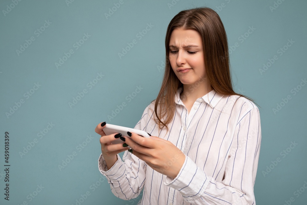 Concentrated attractive young blonde woman wearing casual white shirt isolated over blue background wall holding smartphone and playing online games looking at gadjet screen Concentrated attractive young blonde woman wearing casual white shirt isolated over blue background wall holding smartphone and playing online games looking at gadjet screen
