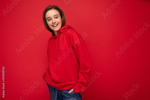 Shot of beautiful happy cool smiling brunette little girl wearing trendy red hoodie standing isolated over red background wall looking at camera © Ivan Traimak