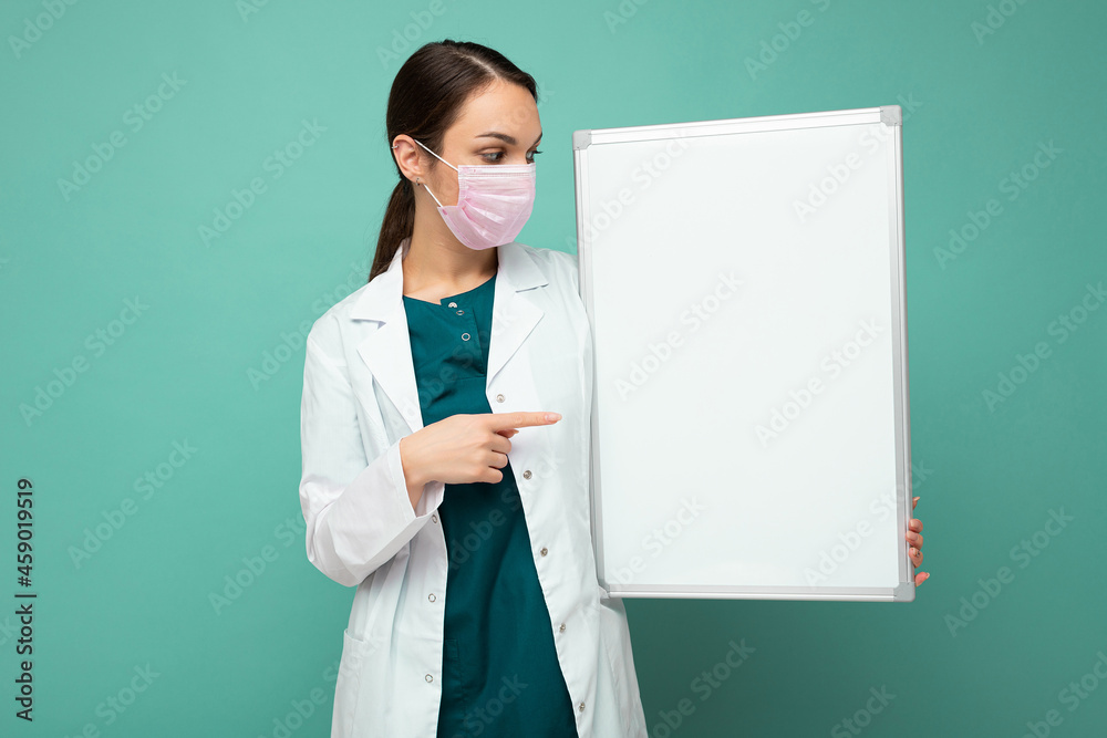 Young attractive nurse in protective face mask and white medical coat holding an empty magnetic board isolated on blue background