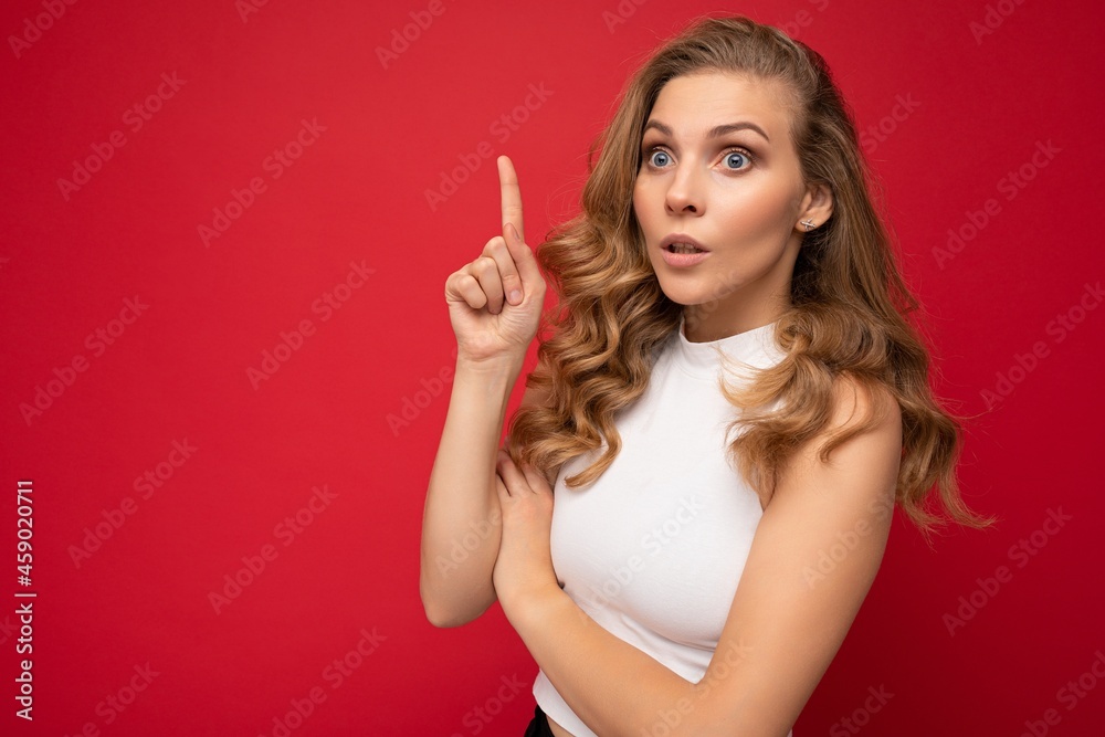 Young beautiful shocked positive blonde curly woman with sincere emotions wearing white top isolated over red background with free space and pointing at copy space for text Young beautiful shocked positive blonde curly woman with sincere emotions wearing white top isolated over red background with free space and pointing at copy space for text