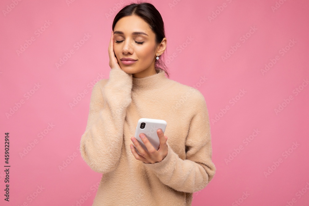 Attractive cute young brunet woman wearing beige warm sweater standing isolated over pink background surfing on the internet via phone and thinking about something pleasant Attractive cute young brunet woman wearing beige warm sweater standing isolated over pink background surfing on the internet via phone and thinking about something pleasant