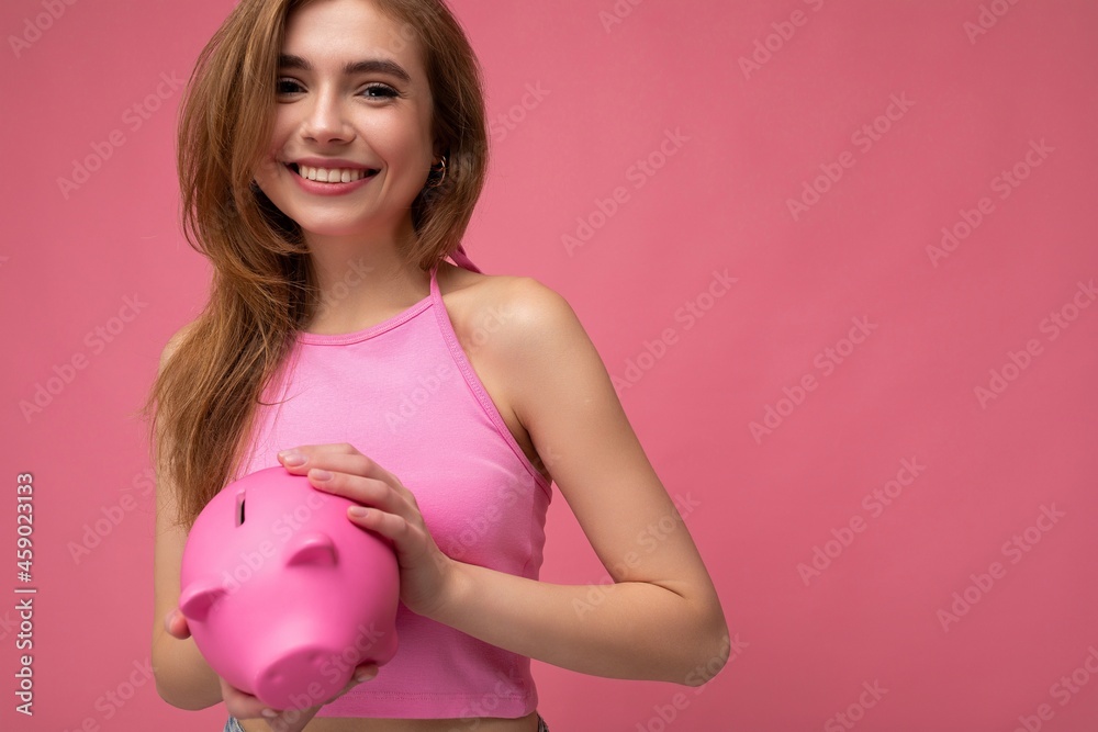 Portrait photo of happy positive smiling young beautiful attractive blonde woman with sincere emotions wearing pink summer top isolated over pink background with copy space and holding pink piggy box Portrait photo of happy positive smiling young beautiful attractive blonde woman with sincere emotions wearing pink summer top isolated over pink background with copy space and holding pink piggy box