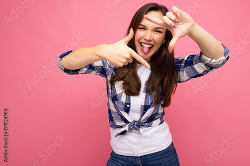 Young positive delightful smiling beautiful brunette woman with sincere emotions wearing hipster check shirt making frame with hands, taking photo with imaginary camera and isolated on pink background