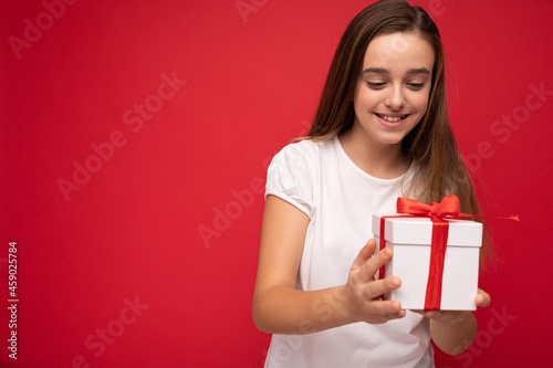 Photo of beautiful positive happy little female child isolated over red bacground wearing casual white t-shirt wall holding gift box and having fun