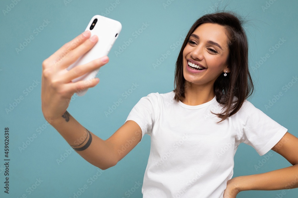 Attractive charming young smiling happy woman holding and using mobile phone taking selfie wearing stylish clothes isolated over wall background Attractive charming young smiling happy woman holding and using mobile phone taking selfie wearing stylish clothes isolated over wall background
