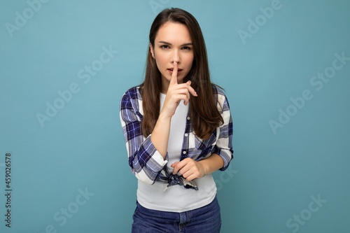 Young serious beautiful brunet woman with sincere emotions wearing trendy check shirt standing isolated on blue background with empty space and showing shhh gesture. Keep secret concept