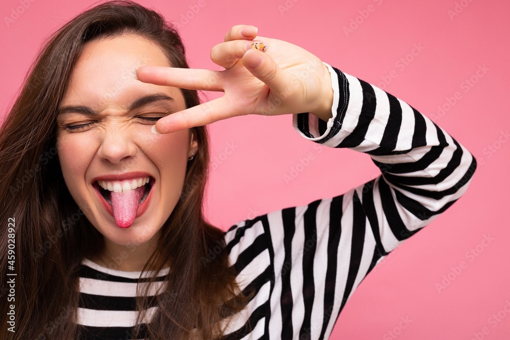 Photo of young european positive smiling winsome happy beautiful brunette woman with sincere emotions wearing casual striped pullover isolated on pink background with copy space and showing tongue and Photo of young european positive smiling winsome happy beautiful brunette woman with sincere emotions wearing casual striped pullover isolated on pink background with copy space and showing tongue and
