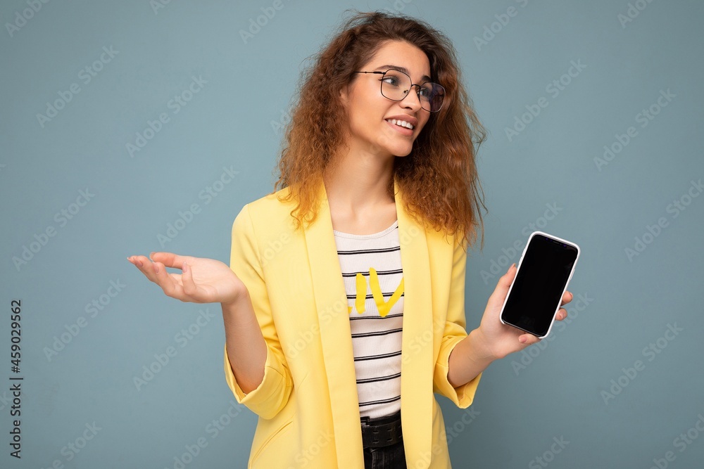 attractive young woman with curly dark blond hair wearing yellow jacket and optical glasses isolated on background holding and showing mobile phone with empty space for cutout looking to the side and attractive young woman with curly dark blond hair wearing yellow jacket and optical glasses isolated on background holding and showing mobile phone with empty space for cutout looking to the side and