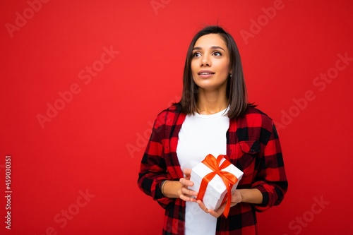Photo shot of pretty positive young brunette woman isolated over red background wall wearing white casual t-shirt and red and black shirt holding white gift box with red ribbon and looking up © Ivan Traimak