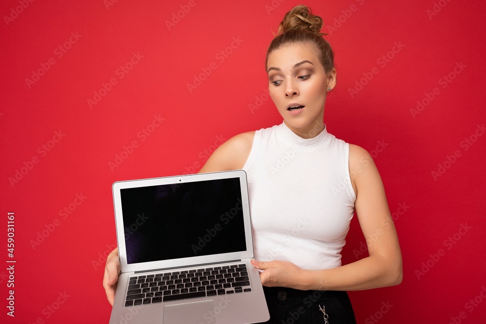 beautiful blond young woman with gathered hair looking down at netbook keyboard holding computer laptop with empty monitor screen with mock up and copy space wearing white t-shirt isolated on red wall beautiful blond young woman with gathered hair looking down at netbook keyboard holding computer laptop with empty monitor screen with mock up and copy space wearing white t-shirt isolated on red wall