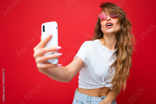 Closeup photo of sexy beautiful adult blonde woman holding mobile phone taking selfie photo using smartphone camera wearing sunglasses everyday stylish outfit isolated over colorful wall background