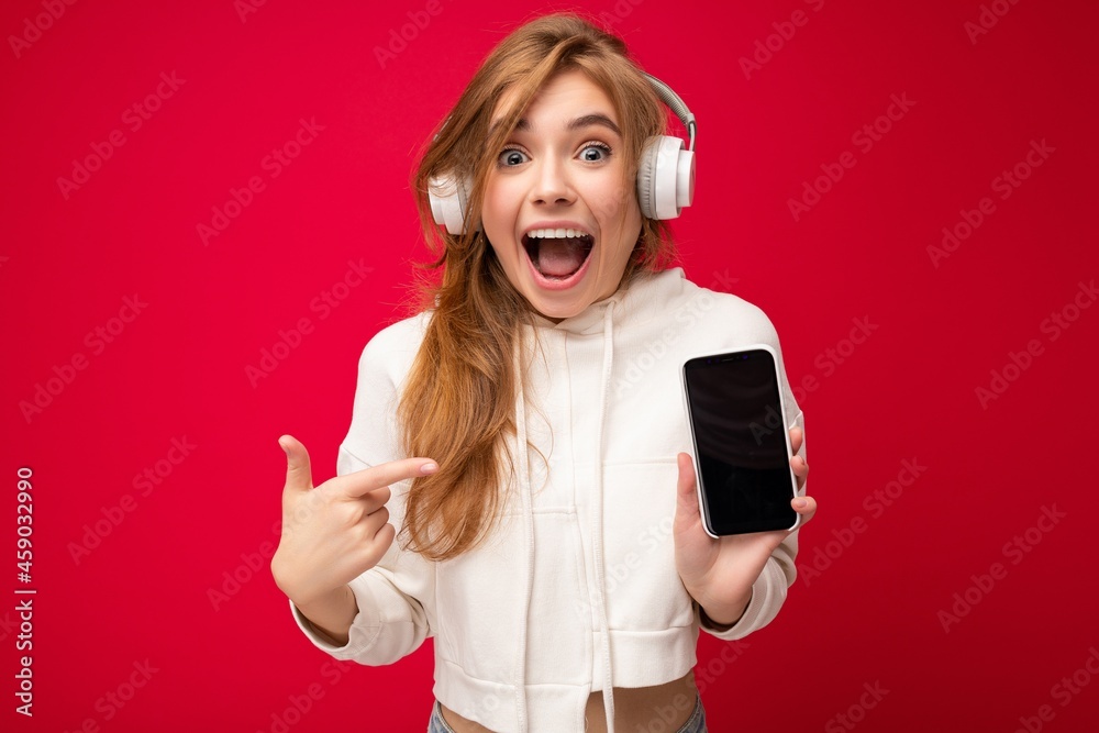 Beautiful positive amazed young woman wearing stylish casual outfit isolated on background wall holding and showing mobile phone with empty display for mockup wearing white bluetooth headphones