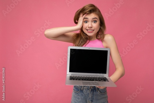 Shot of charming surprised young woman holding computer laptop with empty monitor display wearing pink t-shirt looking at camera isolated on pink wall