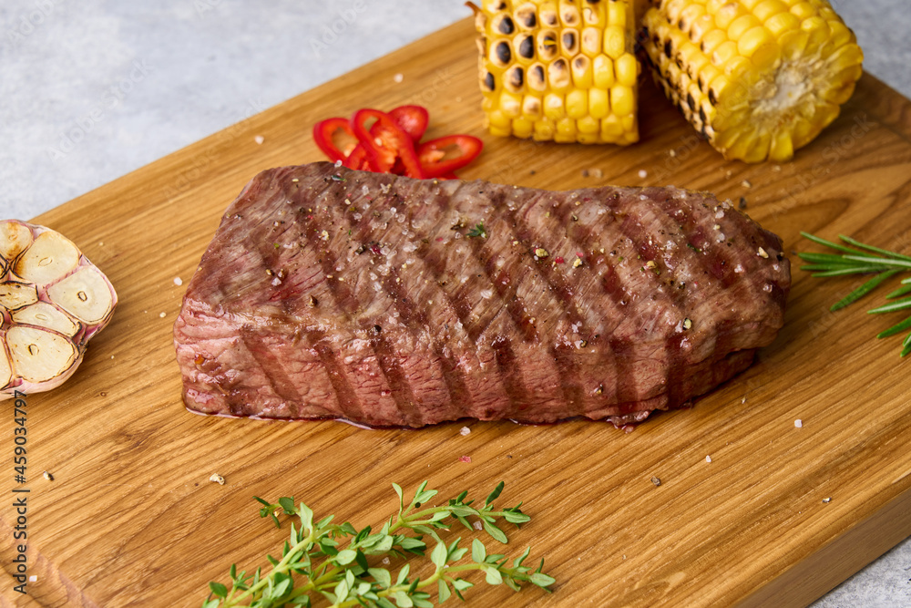 Roasted striploin steak with grilled corn, garlic, red chili pepper, salt, rosemary, thyme. Delicious beef meat with condiment on wooden chopping board. Side view, closeup, horizontal
