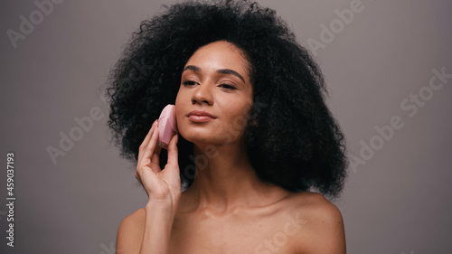 young african american woman with bare shoulders using cleansing device on face isolated on grey