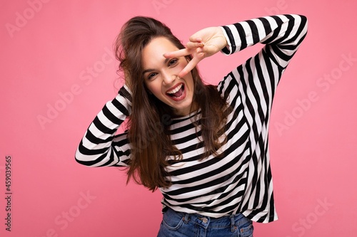 Photo of young european positive smiling winsome joyful happy beautiful brunette woman with sincere emotions wearing casual striped pullover isolated on pink background with copy space