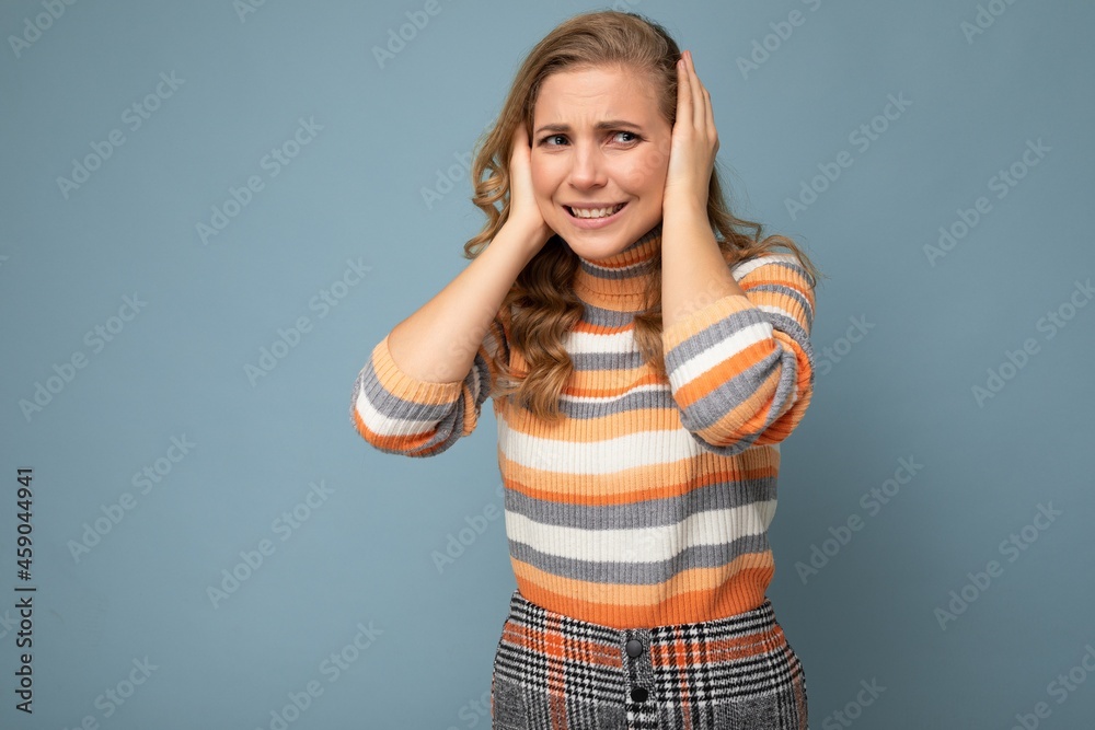 Photo of young pretty nice blonde curly woman with sincere emotions wearing casual striped pullover isolated on blue background with free space and covering ears trying not to hear