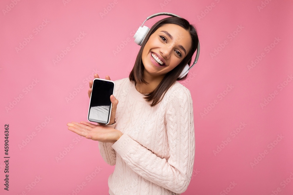 Beautiful happy smiling young woman wearing stylish casual outfit isolated on background wall holding and showing mobile phone with empty display for mockup wearing white bluetooth headphones Beautiful happy smiling young woman wearing stylish casual outfit isolated on background wall holding and showing mobile phone with empty display for mockup wearing white bluetooth headphones