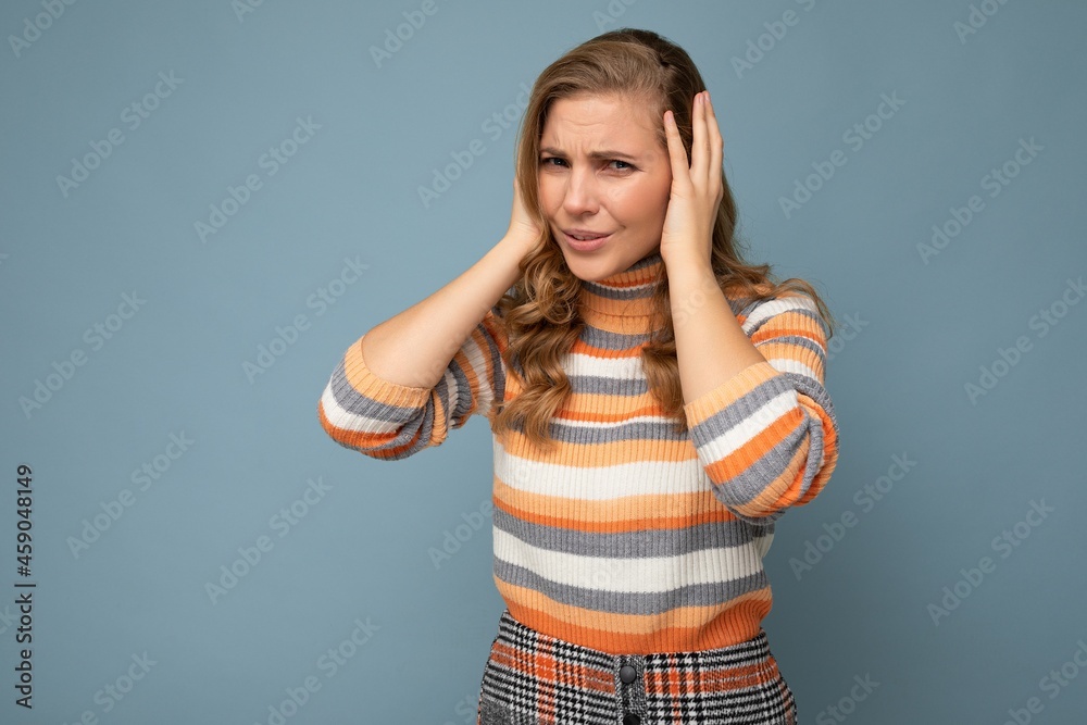 Photo of emotional dissatisfied young attractive blonde curly woman with sincere emotions wearing casual striped pullover isolated on blue background with copy space and covering ears trying not to Photo of emotional dissatisfied young attractive blonde curly woman with sincere emotions wearing casual striped pullover isolated on blue background with copy space and covering ears trying not to