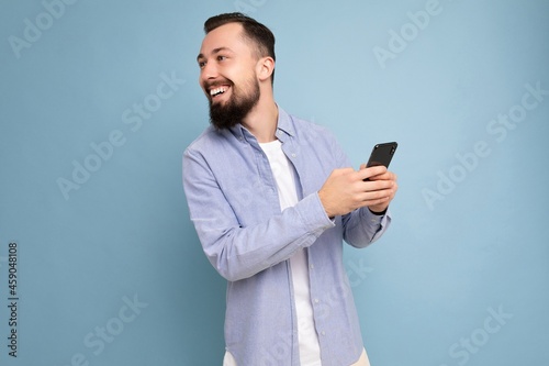 Portrait of smiling handsome young brunette unshaven man with beard wearing stylish white t-shirt and blue shirt isolated over blue background with empty space holding in hand and using phone