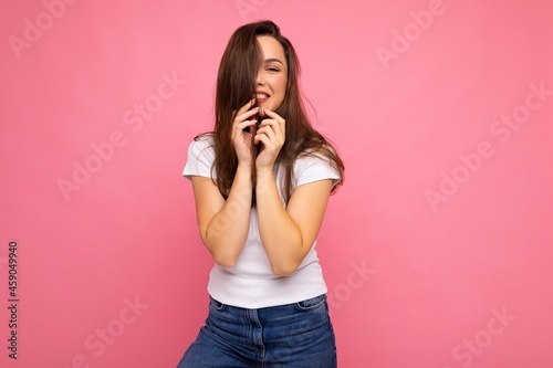 Young beautiful brunet woman. Trendy lady in casual summer white t-shirt for mock up. Positive female shows facial sincere emotions. Funny model isolated on pink background with free space