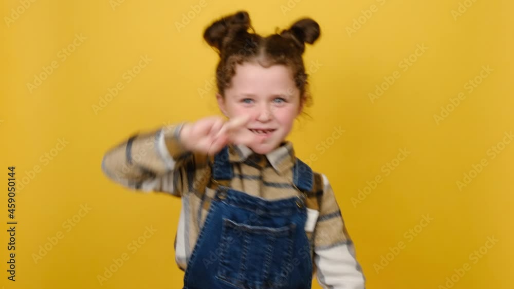 Cheerful funny little caucasian kid girl dancing clenching fists showing victory sign waving hands, posing isolated over yellow color background wall in studio. Childhood emotional lifestyle concept Cheerful funny little caucasian kid girl dancing clenching fists showing victory sign waving hands, posing isolated over yellow color background wall in studio. Childhood emotional lifestyle concept