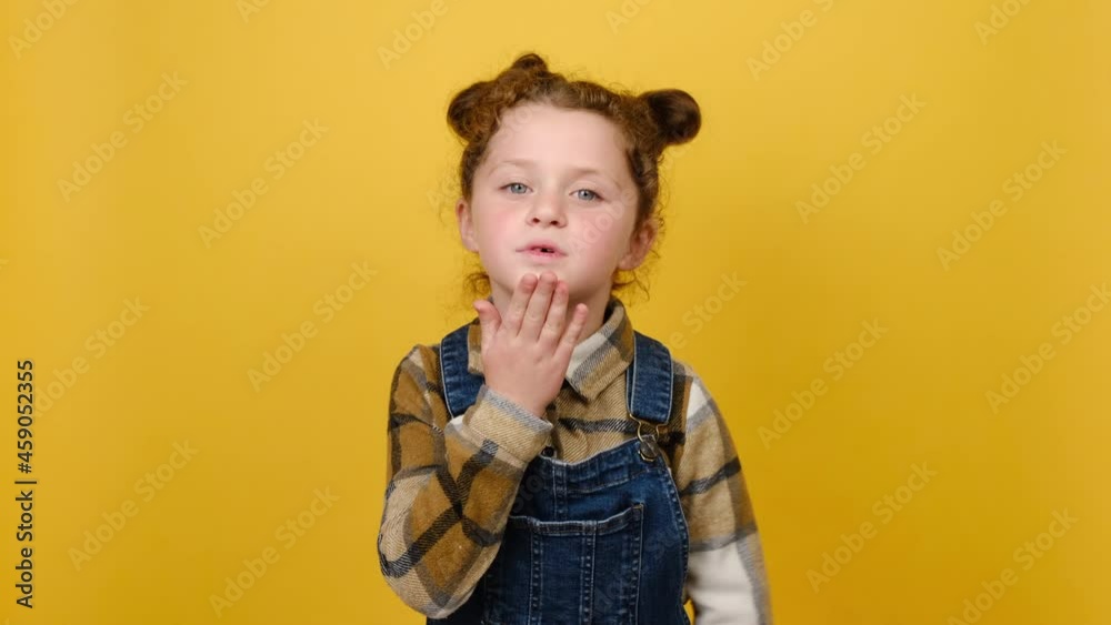 Portrait of cheerful charming pretty little child girl blowing sending air kiss, wears shirt, posing isolated over yellow color wall background in studio. Childhood emotion lifestyle concept Portrait of cheerful charming pretty little child girl blowing sending air kiss, wears shirt, posing isolated over yellow color wall background in studio. Childhood emotion lifestyle concept