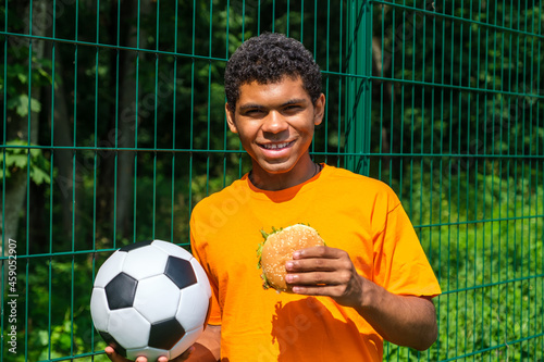 Side view portrait of young African-American man holding soccer ball while standing against fence in sports court outdoors, copy space © Serhii