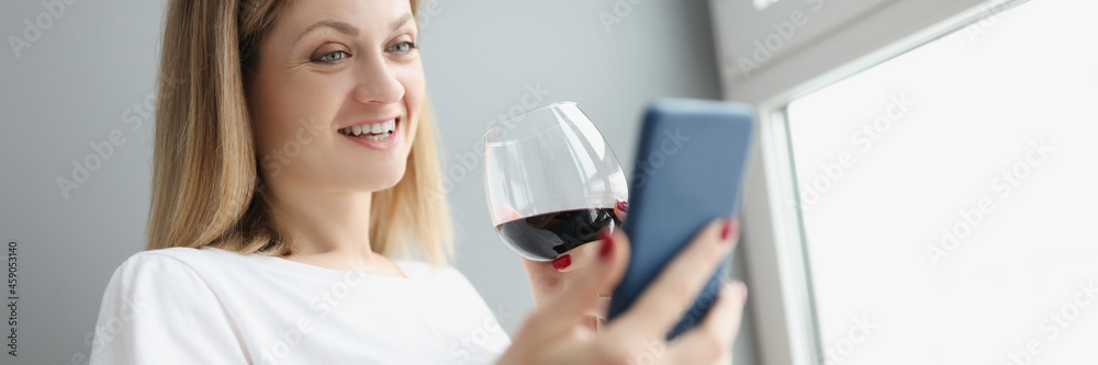 Naklejka premium Young woman holding mobile phone in her hands and drinking red wine from glass at home