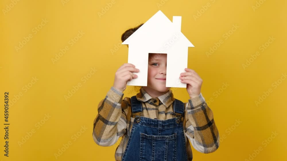 Portrait of pretty smiling caucasian child girl holding small paper white home, friendly looking at camera, posing isolated over yellow studio background. Family house, housing and mortgage concept Portrait of pretty smiling caucasian child girl holding small paper white home, friendly looking at camera, posing isolated over yellow studio background. Family house, housing and mortgage concept