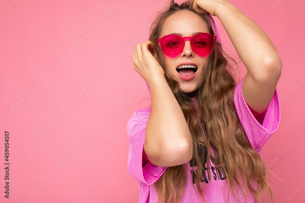 Portrait photo of pretty positive smiling young blonde curly woman isolated over pink background wall wearing casual pink t-shirt and stylish colourful sunglasses looking at camera Portrait photo of pretty positive smiling young blonde curly woman isolated over pink background wall wearing casual pink t-shirt and stylish colourful sunglasses looking at camera