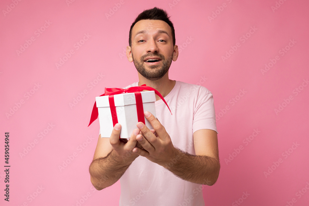 Handsome positive amazing brunette young unshaven man with beard isolated over pink background wall wearing pink t-shirt holding white gift box with red ribbon and looking at camera Handsome positive amazing brunette young unshaven man with beard isolated over pink background wall wearing pink t-shirt holding white gift box with red ribbon and looking at camera
