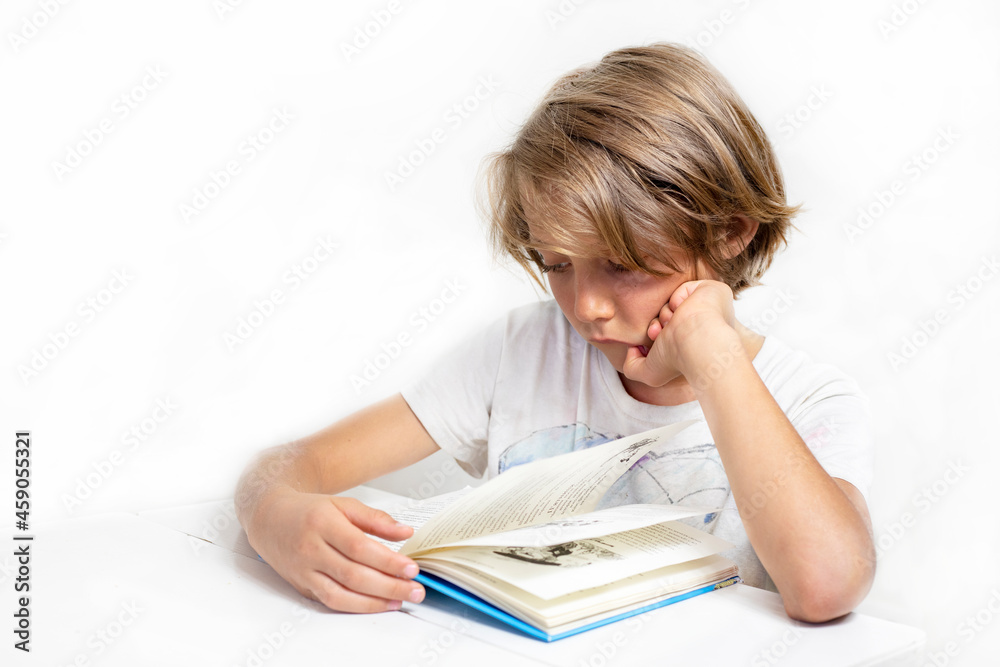 boy reads an interesting book at his desk against the background of a white wall, back to school, education. boy reads an interesting book at his desk against the background of a white wall, back to school, education.