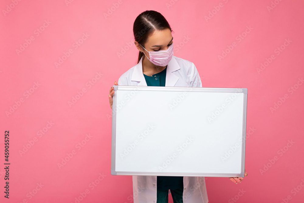 Photo of young attractive nurse in protective face mask holding an empty magnetic board with copy space for information isolated on pink background Photo of young attractive nurse in protective face mask holding an empty magnetic board with copy space for information isolated on pink background