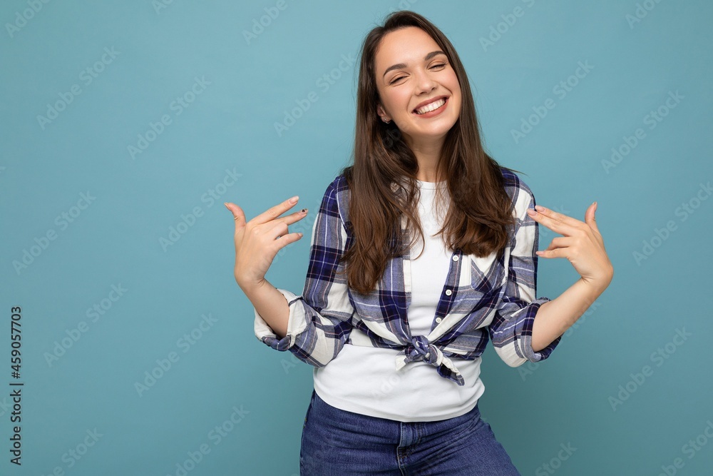 Shot of young positive smiling beautiful brunette woman with sincere emotions wearing trendy check shirt standing isolated on blue background with free space and pointing at yourself