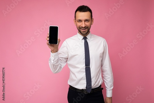 Handsome happy cool young man good looking wearing casual stylish clothes standing isolated over colourful background wall holding smartphone and showing phone with empty screen display looking at