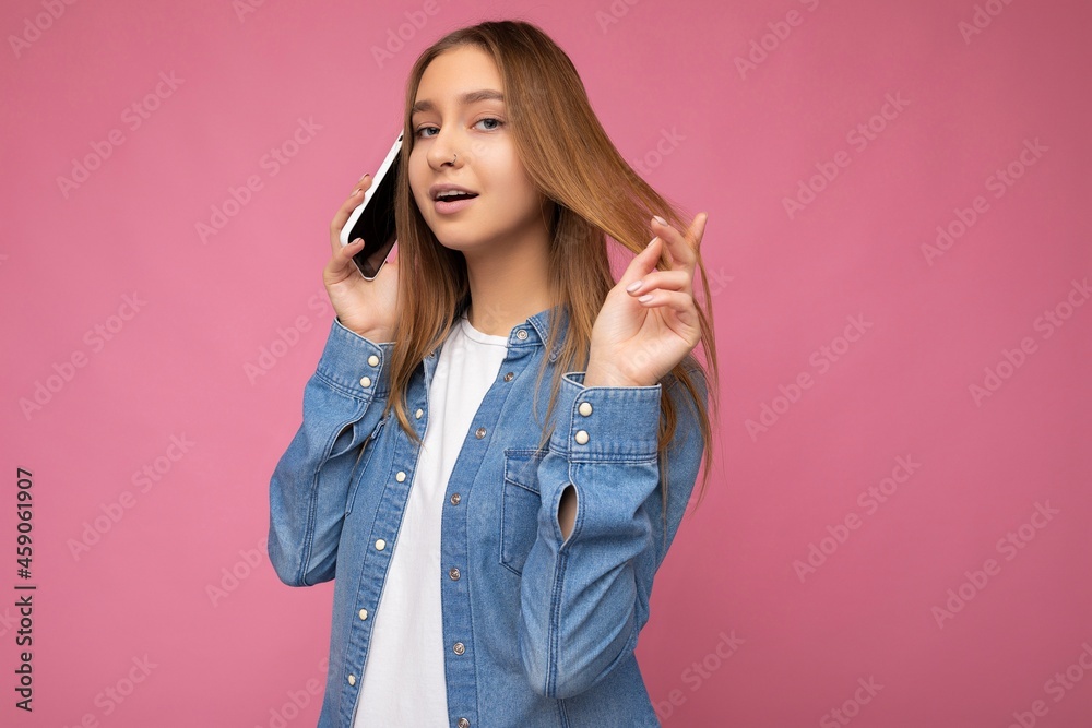 Photo of sexy beautiful happy young blonde woman wearing casual blue jean shirt isolated over pink background holding in hand and talking on mobile phone with boyfriend and flirting looking at camera Photo of sexy beautiful happy young blonde woman wearing casual blue jean shirt isolated over pink background holding in hand and talking on mobile phone with boyfriend and flirting looking at camera