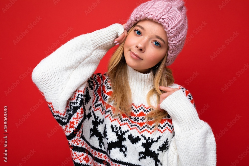 Shot of charming fascinating pretty young blonde woman isolated over red background wall wearing winter sweater and pink hat looking at camera Shot of charming fascinating pretty young blonde woman isolated over red background wall wearing winter sweater and pink hat looking at camera