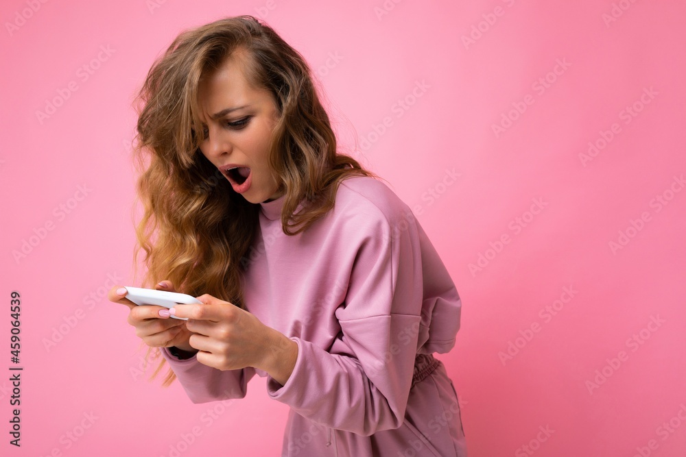 Side-profile amazed surprised blonde young woman with open mouth wearing pink hoodie isolated over pink background with copy space holding and playing games via telephone looking at device display Side-profile amazed surprised blonde young woman with open mouth wearing pink hoodie isolated over pink background with copy space holding and playing games via telephone looking at device display