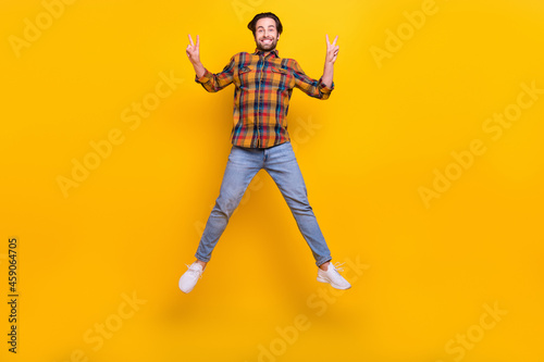 Full body photo of crazy brunet young guy jump show v-sign wear shirt jeans sneakers isolated on yellow background