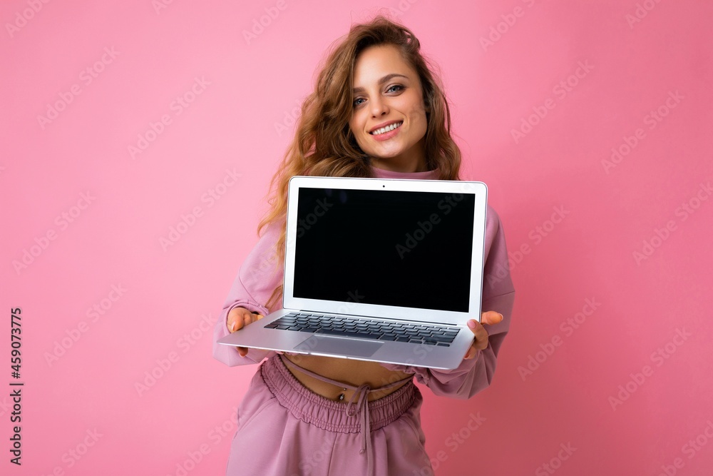Photo of beautiful charming happy smiling dark blond curly young woman lookind at camera holding computer laptop with empty monitor screen with mock up and copy space wearing pink hoodie isolated over Photo of beautiful charming happy smiling dark blond curly young woman lookind at camera holding computer laptop with empty monitor screen with mock up and copy space wearing pink hoodie isolated over