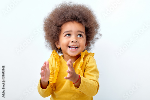 Afro fluffy child girl clapping and applauding, happy and joyful, smiling. Adorable charming kid in yellow shirt have fun isolated on white studio background, looking up. Portrait, copy space.