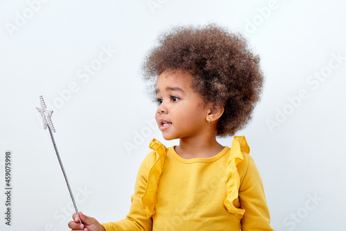 Cute african american girl wth curly fluffy hair holding magic wand, isolated on white studio wall. Funny little black girl wearing yellow casual shirt. Childhood lifestyle concept photo